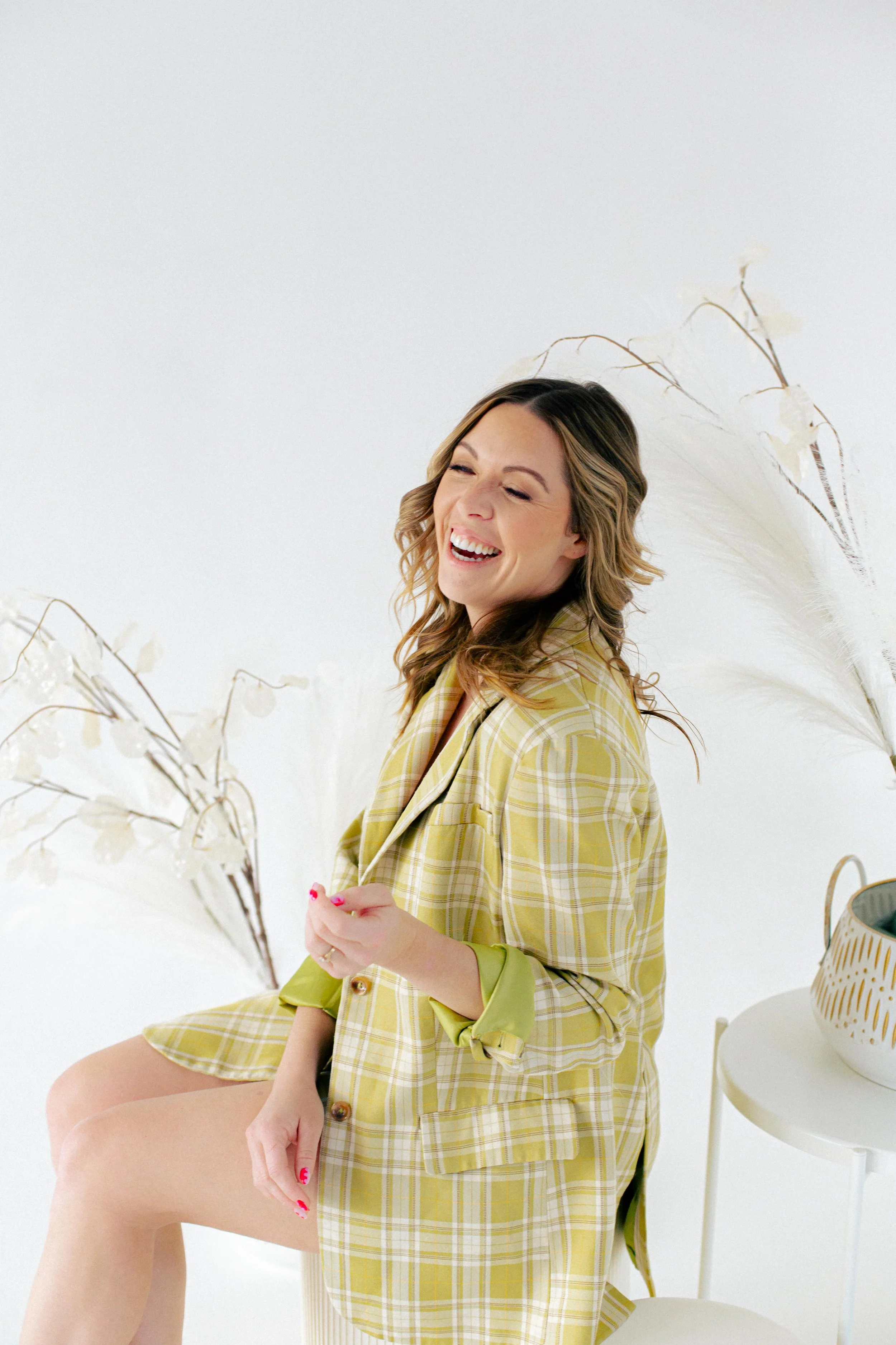 Gabby Pinkerton laughing on a stool against a white backdrop, in a yellow plaid blazer